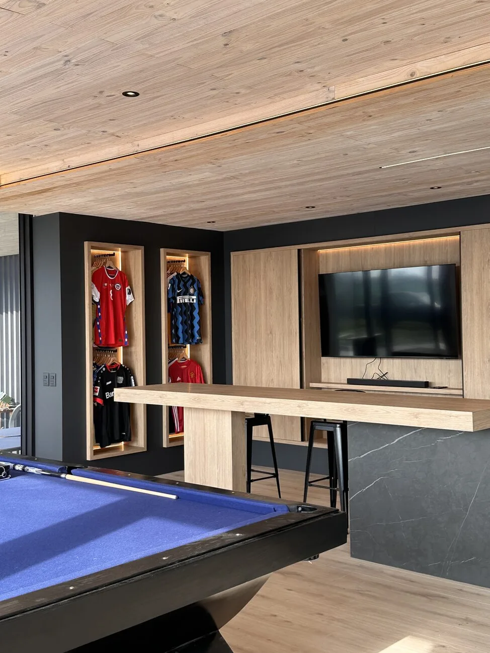 Barra alta de madera frente al mueble de televisión de roble, con nichos verticales iluminados y una mesa de pool en primer plano