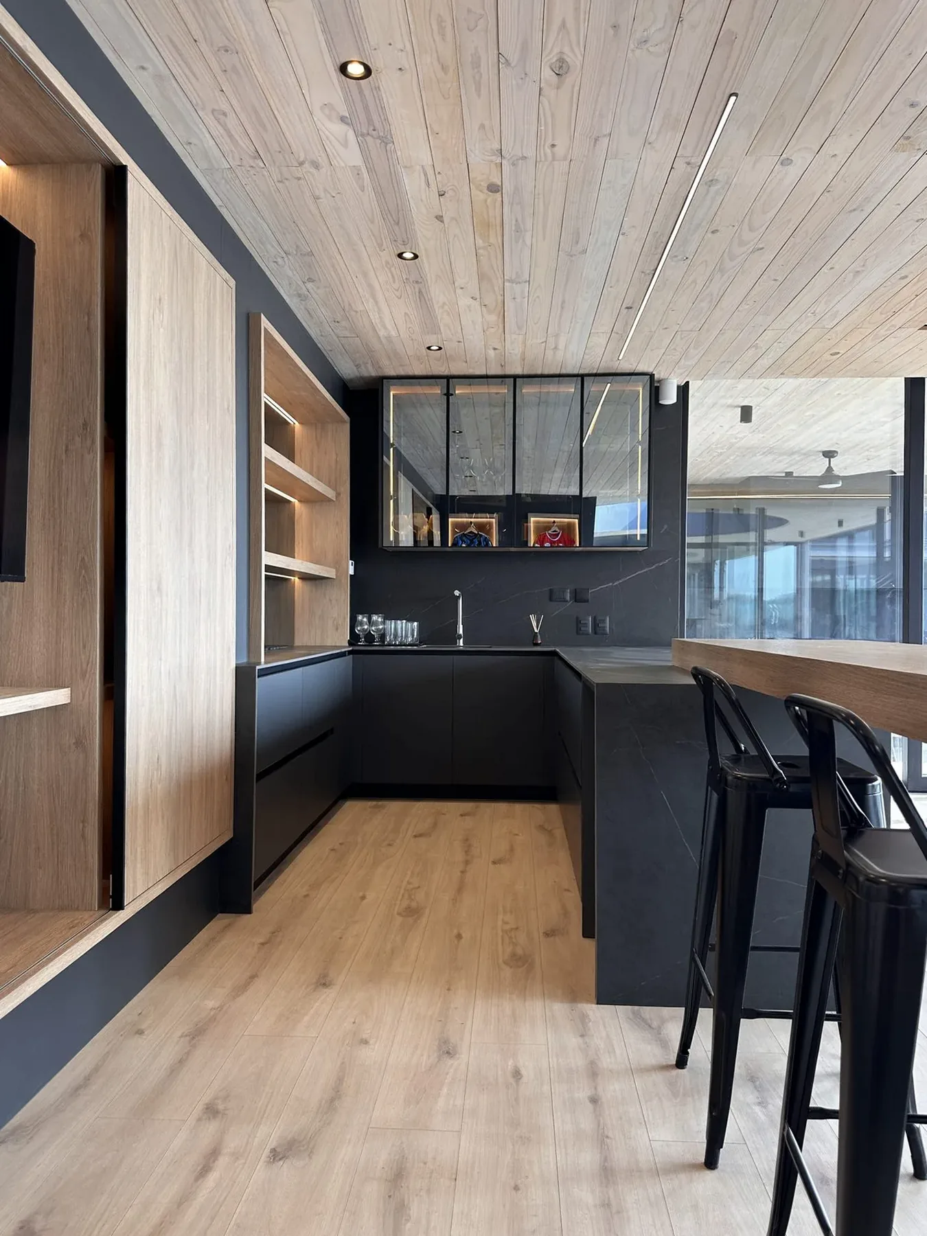 Cocina en U negro mate con cubierta de porcelanato grafito, vitrinas de perfilería negra iluminadas y barra alta de madera bajo un cielo entablado