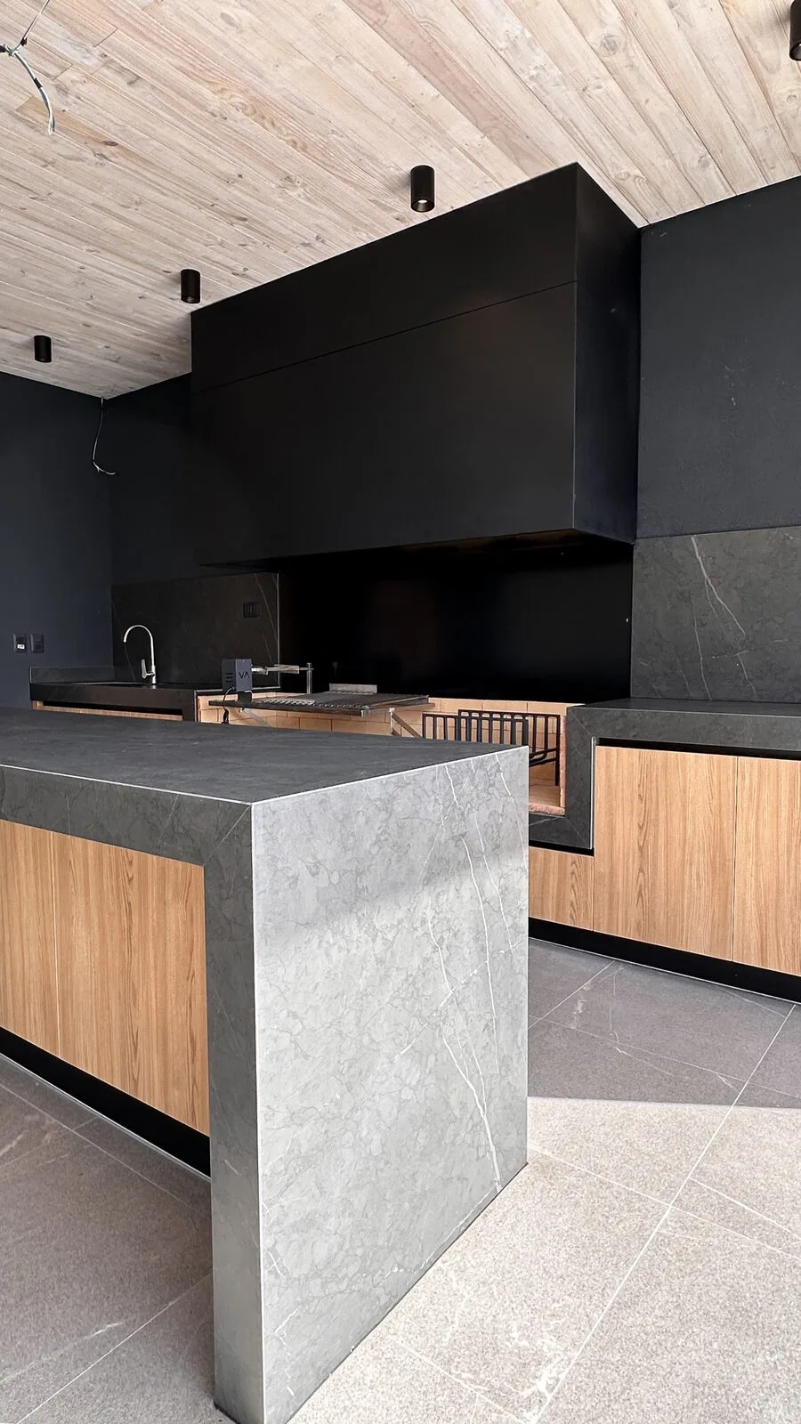 Quincho con parrilla bajo una campana negra, isla de porcelanato veteado y muebles de roble, con cielo entablado